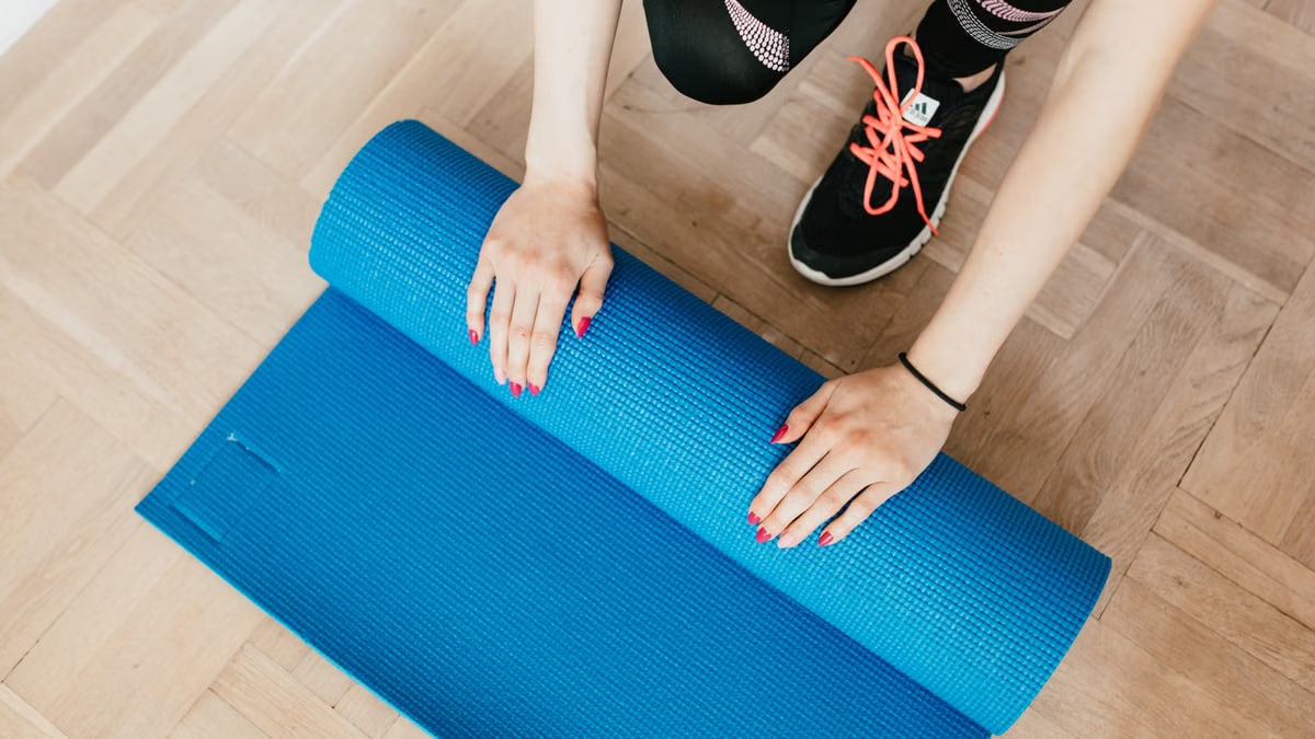 From above of unrecognizable female athlete in leggings and sneakers unfolding sport mat for practicing yoga in morning at home